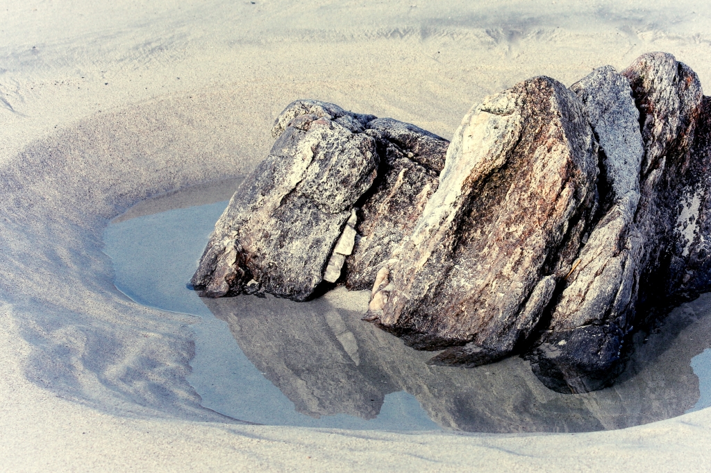 Photo of a granite rock & its reflection in a tide pool in pale sand. 