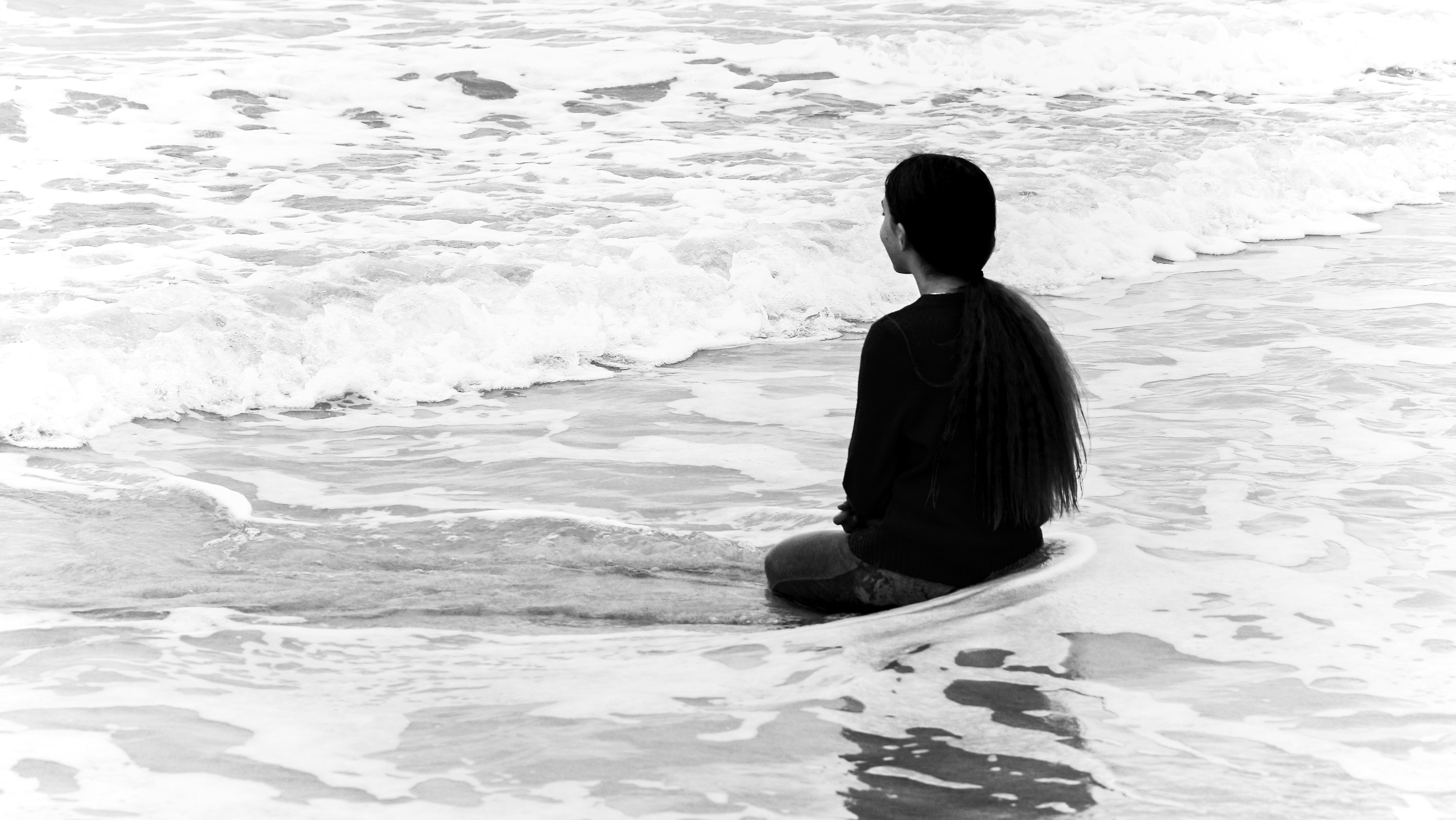 Black and white photograph of a girl kneeling in the surf, her back to the camera. 