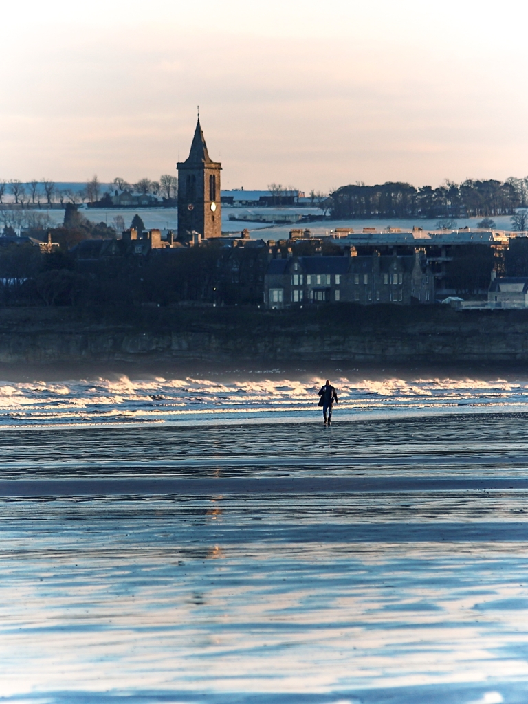 Photo looking back at St. Andrews along West Sands beach with white surf and wet sand, and a lone figure in the distance. 