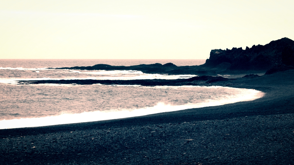 sepia tinted photo of a black sand beach, showing two bays, some rocks and bright white surf line. 