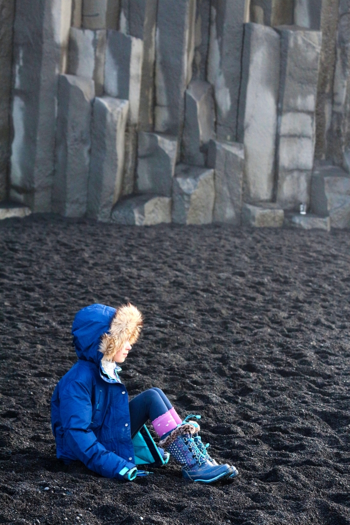 Photo of a small human in blue winter coat, blue leggins and blue snow boots sitting on a black sand beach with black basalt columns in the background. 