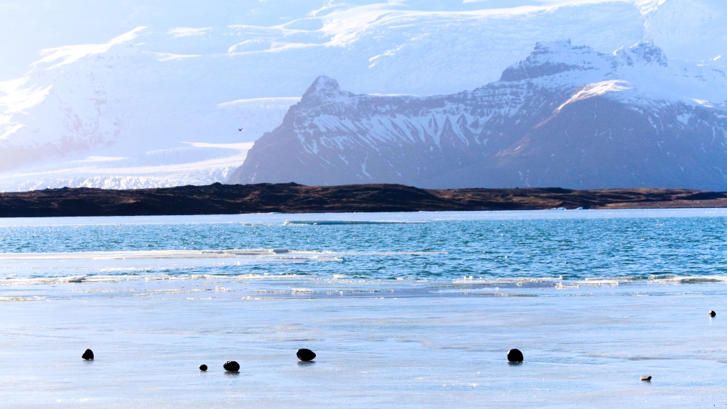 Photo of half frozen lagoon, distant snowy mountains and a glacier. 