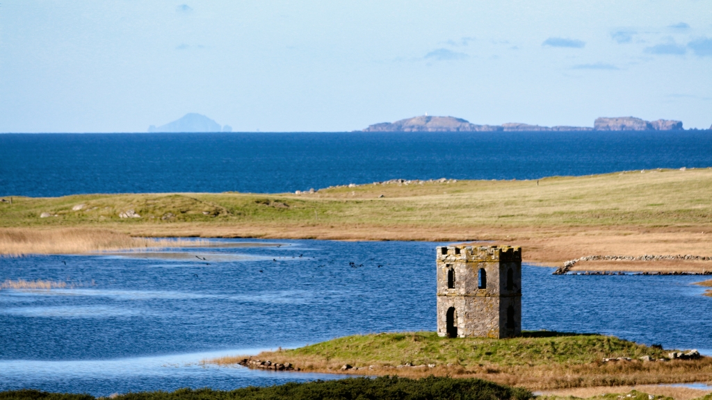 Photo looking out towards St Kilda from North Uist, in the foreground there's a loch with a wee ruined tower on an old broch island. 