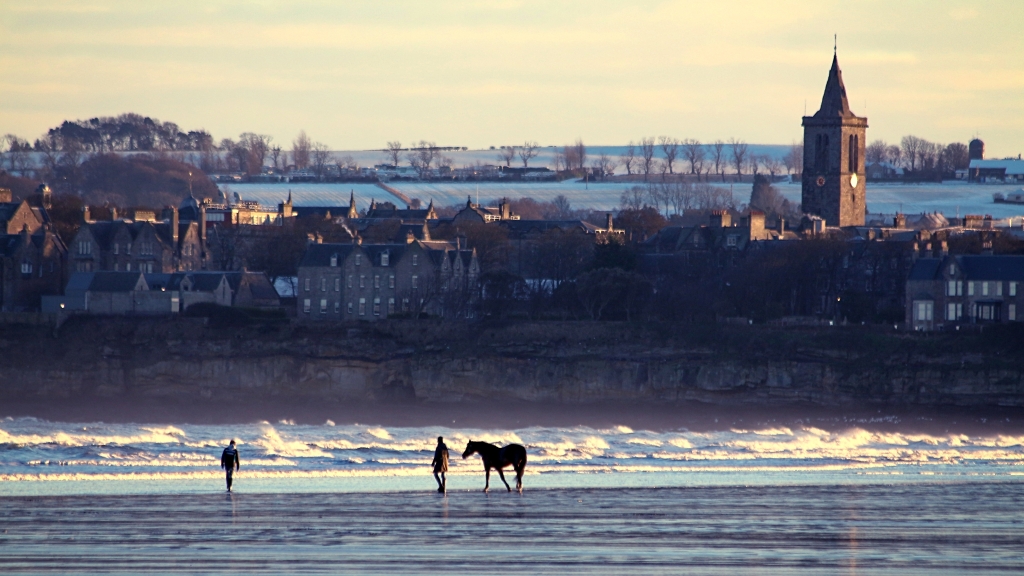 Shot from the beach looking towards St. Andrews. There's a bloke with a horse on the beach & one of the university towers on the skyline, snow on the hill behind and wintery light. 